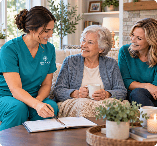 Caregiver in a teal True North Home Care top writing in a notebook beside an older woman holding a mug and a younger woman on a sofa