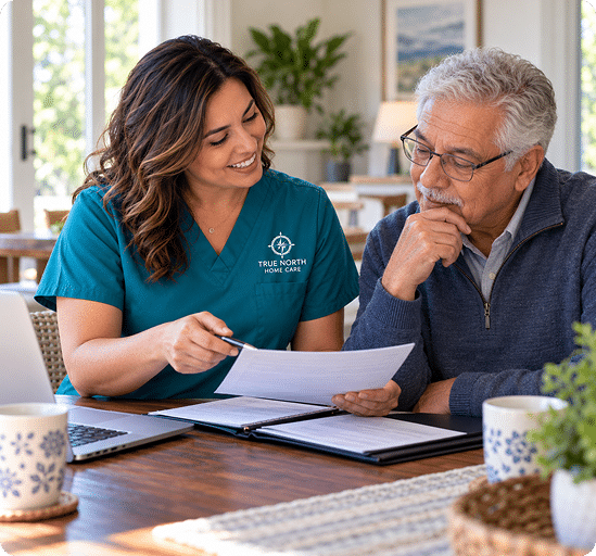 Caregiver in a teal True North Home Care top going over paperwork at a kitchen table with an older man in glasses